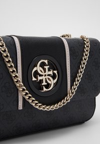 Černá kabelka s texturou a zlatým řetízkem přes rameno. Obsahuje kulatý ozdobný prvek s logem, světle růžové detaily a ražené vzory na materiálu.