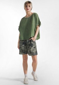 Junge Person steht, trägt ein locker sitzendes grünes Oberteil, gemusterte Shorts, weiße Socken und Turnschuhe vor weißem Hintergrund.