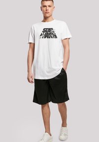 Hvit bomull T-skjorte med en svart "STAR WARS" grafikk, kombinert med svarte treningsshorts og hvite joggesko. Glatt tekstur, uformell passform.