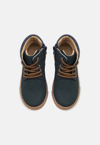 Tmavě modré semišové kotníkové boty s hnědými šňůrkami, bočními zipy a béžovými gumovými podrážkami; hladký povrch s minimálními akcenty.