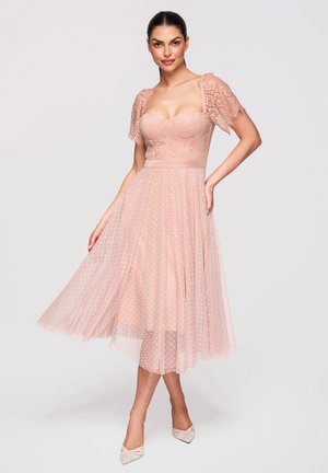 Femme posant dans une robe midi rose poudré avec des manches courtes en dentelle et une jupe transparente à pois, portant des escarpins à bout pointu sur un fond blanc.