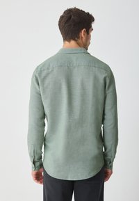 Men's long-sleeve shirt in light green linen, featuring a rounded hem, buttoned cuffs, and a collar. Textured fabric adds subtle depth.