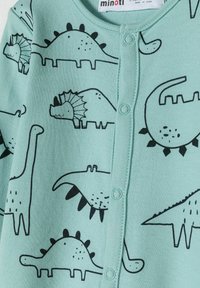 Cárdigan de algodón en verde claro con ilustraciones de dinosaurios negros, cierre de botones y cuello acanalado. Diseño sencillo con un patrón lúdico.