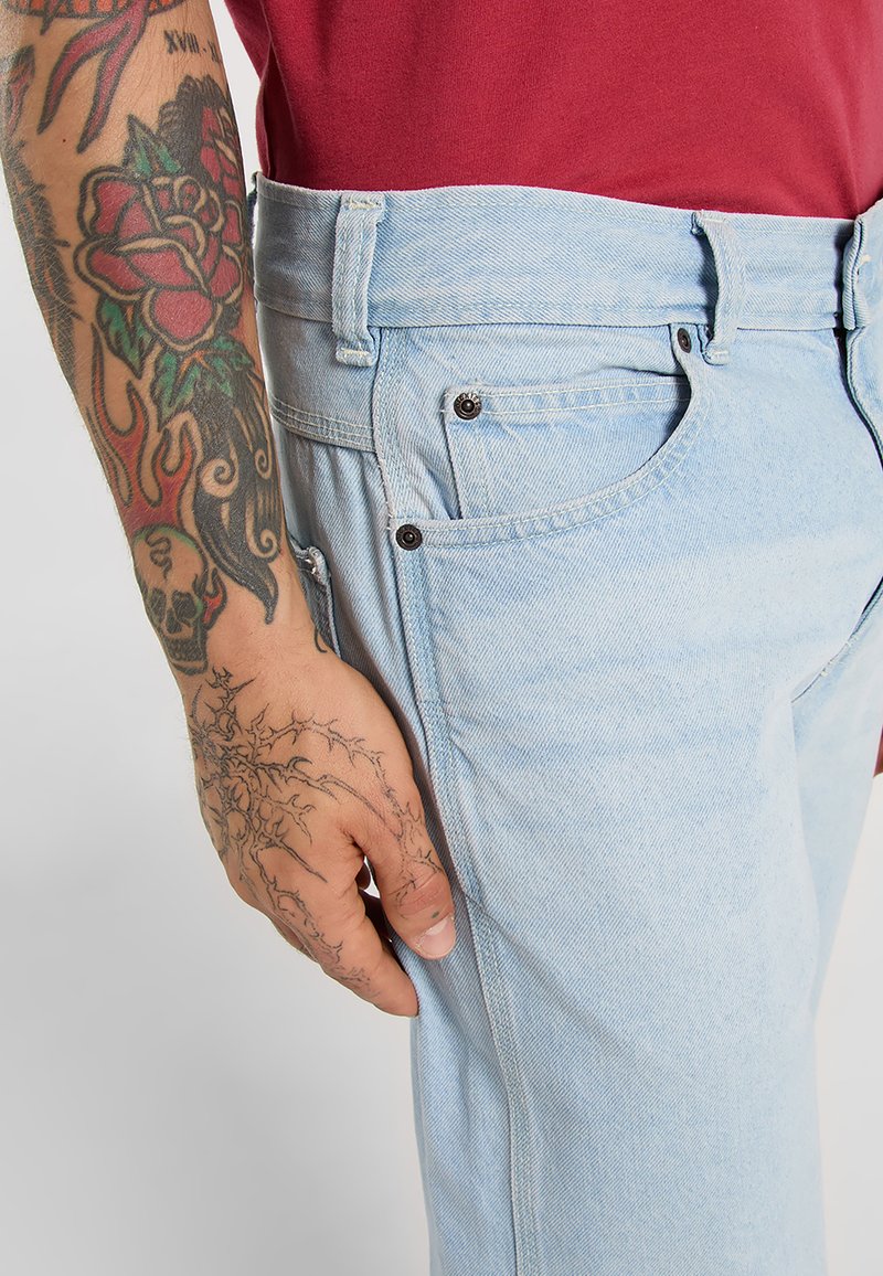 Getatoeëerde arm en hand naast een lichtblauwe denim jeanszak, met een rood shirt in de taille gestopt.