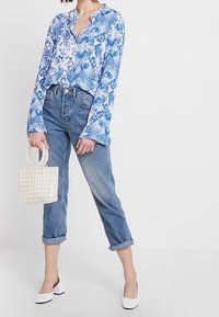 Blusa a maniche lunghe con motivo floreale blu, jeans azzurri, scarpe bianche con tacco a blocco e una piccola borsa a tracolla bianca con perline.