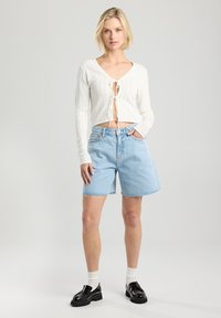 Valkoinen kevyt neulecardigan, jossa on solmimisvaihtoehto, yhdistettynä vaaleansinisiin farkkushortseihin. Mallilla on päällään mustat loafers-kengät ja valkoiset nilkkasukat.