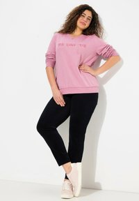 Kvinna med lockigt hår som bär en rosa "Be Unique" sweatshirt, svarta byxor och ljusrosa sneakers, stående med ena handen på höften.