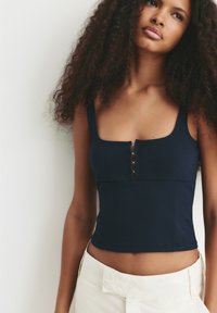 Vrouw met krullend haar in een marineblauwe mouwloze cropped top en hoge witte broek, staande tegen een plain witte achtergrond.