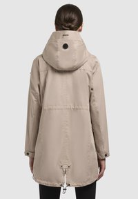 Giacca impermeabile beige con cappuccio, vita regolabile e polsini con bottone. Presenta un logo sul retro e una superficie liscia e opaca.