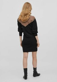 Schwarzes Strickkleid mit braunem Akzent an der Schulter, langen Ärmeln, taillierter Silhouette und einem V-förmigen Detail am Rücken. Schwarze Knöchelstiefel.