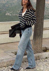 Zwart-wit gestreepte hoodie, beige top, lichtblauwe wijde jeans, zwarte puntige schoenen; staat naast een boom in een buitenomgeving.