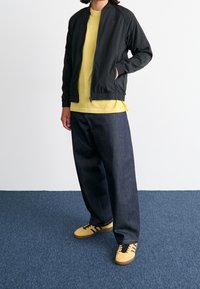 Svart lätt bomberjacka, gul t-shirt, mörkblå oversized jeans och gula sneakers med svarta ränder. Enkel bakgrund och blå matta.