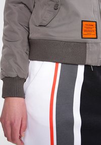 Graue Bomberjacke mit einem gerippten braunen Bündchen, verziert mit einem Logopatch und einer Seitentasche. Kombiniert mit einem weißen Rock, der rote und graue Streifen hat.
