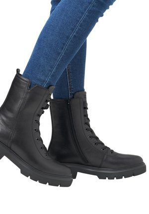 Mustat nahkaiset nilkkurit, joissa sivuvetoketju ja nauhat, yhdistetty sinisiin kapeisiin farkkuihin; kengissä näkyy kestävä pohja ja sileä viimeistely.