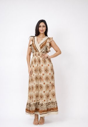 Kobieta stojąca z ręką na biodrze, ubrana w beżową sukienkę maxi z brązowymi kwiatowymi wzorami i marszczonymi rękawami na tle jednolitego białego tła.