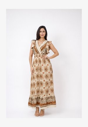 Vrouw staat met hand op heup, draagt een beige maxi-jurk met bruine bloemmotieven en gerimpelde mouwen, tegen een effen witte achtergrond.