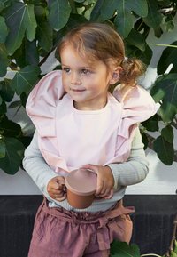 Vaaleanpunainen silikoni-tyyny, jossa on röyhelöreunat, käytetty harmaan pitkänhihaisen paidan päällä. Lapsi pitää ruskeaa kuppia kädessään seisten lehvätaustan edessä.