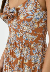Brun blomsterkjole med blå og gule mønstre. Har en sløjfedetalje ved halsen og draperinger i taljen for tekstur.