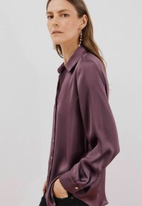 Camicia button-up in seta di un profondo colore viola, con colletto, maniche lunghe e polsini abbottonati. Il tessuto ha una consistenza liscia e una leggera lucentezza.