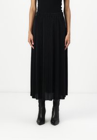 Jupe maxi plissée noire en tissu texturé avec taille élastique, portée avec des bottines noires ajustées.