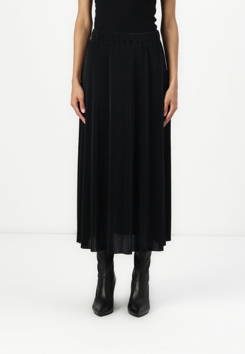 Jupe maxi plissée noire en tissu texturé avec taille élastique, portée avec des bottines noires ajustées.