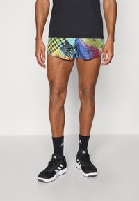 Bunte gemusterte Badeshorts mit abstrakten Designs in Grün, Blau und Gelb. Kombiniert mit schwarzen Sportstrümpfen und Schuhen.