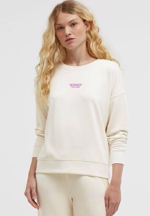 Frau mit langen lockigen blonden Haaren, die einen elfenbeinfarbenen Pullover und passende Hose mit lila "senses the label" Schrift auf der Brust trägt.