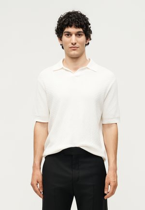 Jeune homme aux cheveux bouclés foncés, portant une chemise blanche à manches courtes avec col et un pantalon noir, debout devant un fond uni.