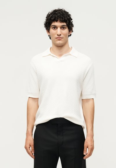 Jeune homme aux cheveux bouclés foncés, portant une chemise blanche à manches courtes avec col et un pantalon noir, debout devant un fond uni.