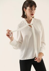 White blouse with a tie, made of smooth fabric. Features a V-neck and long sleeves, paired with black pants. model holding the tie.