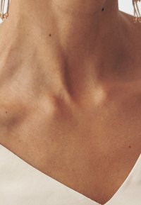 Top z dekoltem w serek w miękkim białym materiale, eksponujący gładką szyję i ramiona z subtelną strukturą skóry oraz lekkimi piegami.