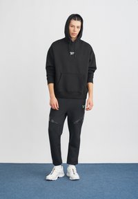Felpa nera con cappuccio e tasca frontale, impreziosita da un logo, abbinata a pantaloni jogger neri a gamba affusolata con branding verde e dettagli in rete. Sneakers bianche.