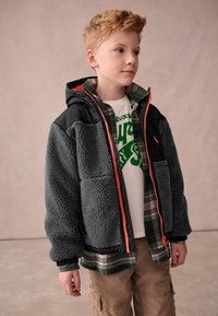 Young boy with short ginger hair wearing a gray fleece jacket over a green plaid shirt and white graphic tee, standing facing slightly right.