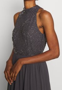 Ärmlösa grå klänning med hög krage. Pärlmönster i en solstråle-design längs livet, flödande kjol i tyg.