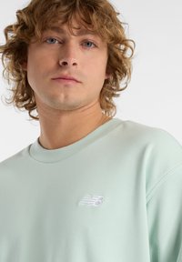 Ljus mintgrön sweatshirt med ribbat rundhals, som har en liten vit broderad New Balance-logotyp på vänster bröst. Mjuk textur.