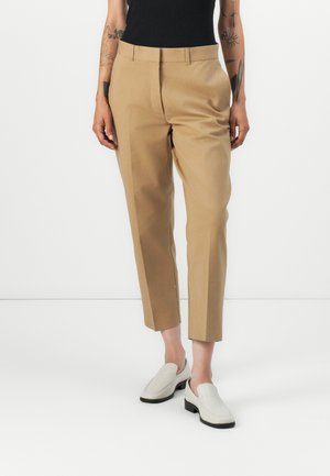 Tan tailored trousers with a high waist, tapered fit, and smooth texture, paired with off-white slip-on shoes.