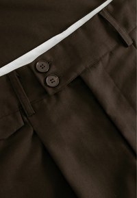 Smeđe krojene hlače s dva tamna gumba, vidljivim pregibima i glatkom teksturom. Imaju bijeli akcent na pojasu.