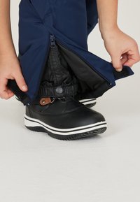 Svarta vattentäta stövlar med vita sulor, kombinerade med marinblå vattentäta byxor. Byxorna har dragkedjor och justerbara muddar för skydd och passform.