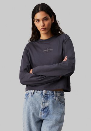 Maglietta a maniche lunghe cropped grigio scuro con logo bianco sul petto, abbinata a jeans azzurri chiari a vita alta con un leggero sbiadimento.