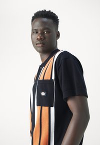 Camisa polo de punto negra con rayas verticales naranjas y blancas, mangas cortas y un bolsillo frontal con una etiqueta.