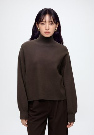 Jeune femme aux longs cheveux foncés porte un pull à col roulé marron foncé et un pantalon assorti, debout devant un fond clair uni.