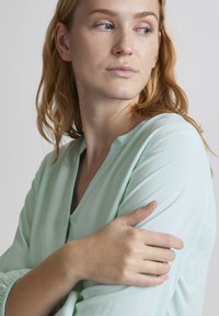 Bluză de culoare verde mentă deschisă, cu mâneci lungi, decolteu în formă de V și manșete plisate. Hingă moale cu o textură fină. Model cu păr ondulat.
