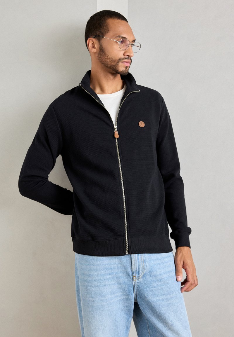 Hombre vestido con chaqueta negra con cremallera sobre una camisa blanca y jeans de color azul claro, de pie contra una pared gris claro lisa, mirando hacia la derecha.