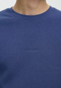 Mėlynas medvilninis džemperis su apvalia iškirpte, kuriame yra iškilmingas "Selected" logotipas ant krūtinės ir lygus paviršius visame džemperi.