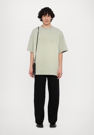 T-shirt surdimensionné vert clair avec texte en relief, associé à un pantalon noir à jambes larges et des chaussures noires. Comprend un petit sac noir à bandoulière.
