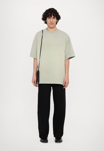 Ανοιχτό πράσινο oversized μπλούζα με ανάγλυφο κείμενο, συνδυασμένη με μαύρο παντελόνι σε ίσια γραμμή και μαύρα παπούτσια. Περιλαμβάνει μια μικρή μαύρη τσάντα ώμου.