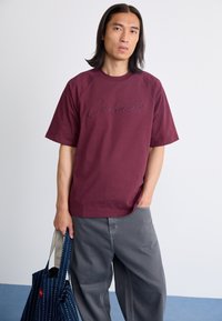 Burgundi rövid ujjú póló, rajta a "Carhartt" logóval, szürke, bő szárú farmernadrággal párosítva. A modellen egy kék mintás vászontáska van.
