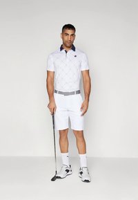 Biele krátke rukávy polo tričko s diamantovým vzorom, námornícka modrá golier, biele šortky, sivý opasok a biele golfové top�ánky. Stojí s golfovou palicou.