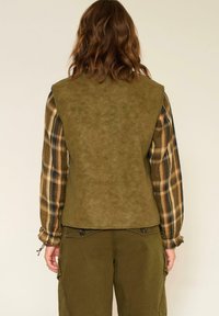 Gilet en daim vert olive avec une texture lisse, présentant un design au dos. Porté par-dessus une chemise à manches longues à carreaux dans des tons terre et un pantalon sombre.