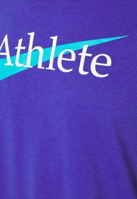 Camiseta de algodón morada con texto blanco que dice "Atleta" junto a un diseño de franjas diagonales azules. Textura suave, estilo casual.